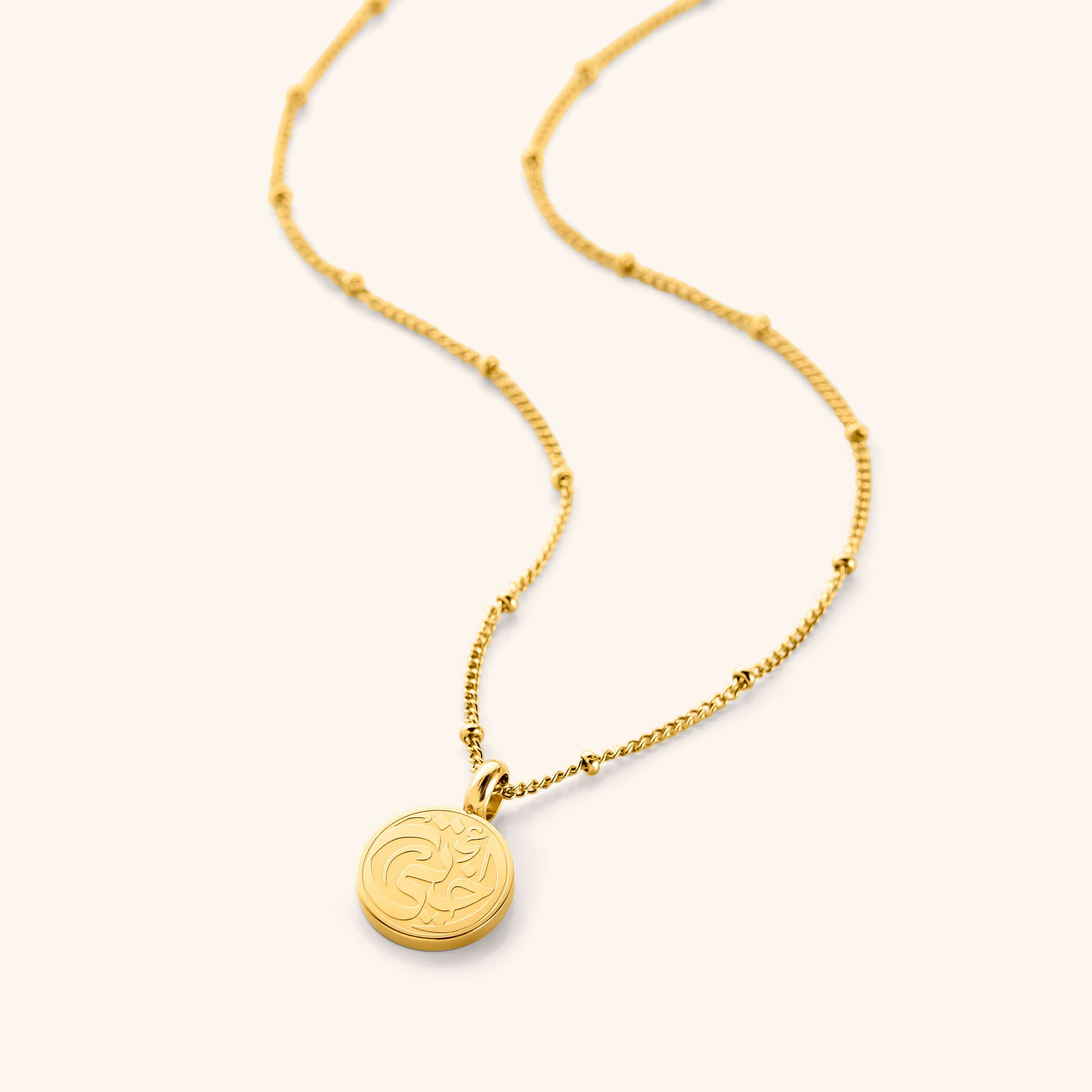 Close-up of Arabic gold pendant with ‘my sister’ inscription on dotted chain, laying flat on velvet pouch